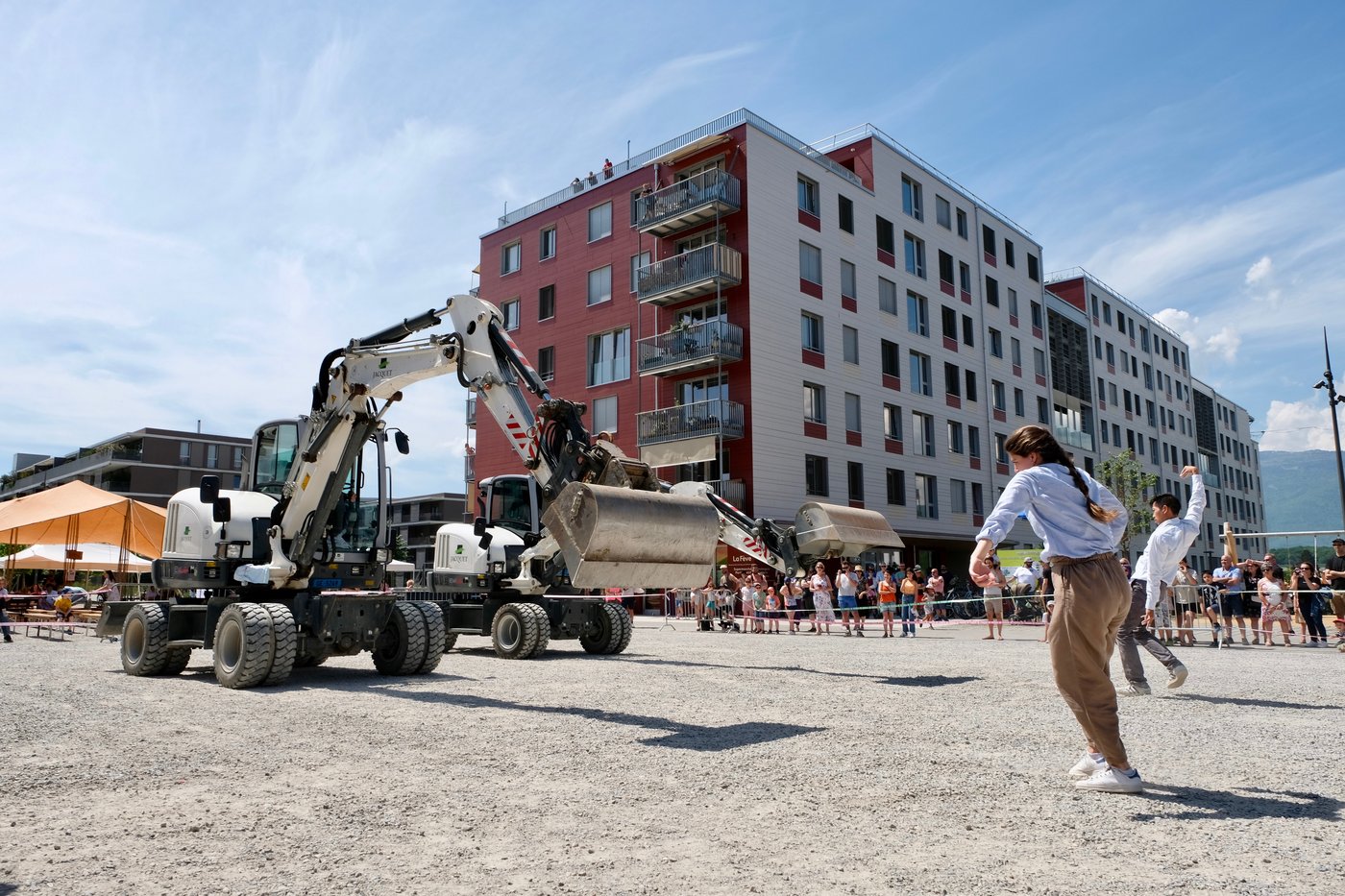 Deux pelleteuses face à deux danseurs sur un terrain de gravier, devant un public