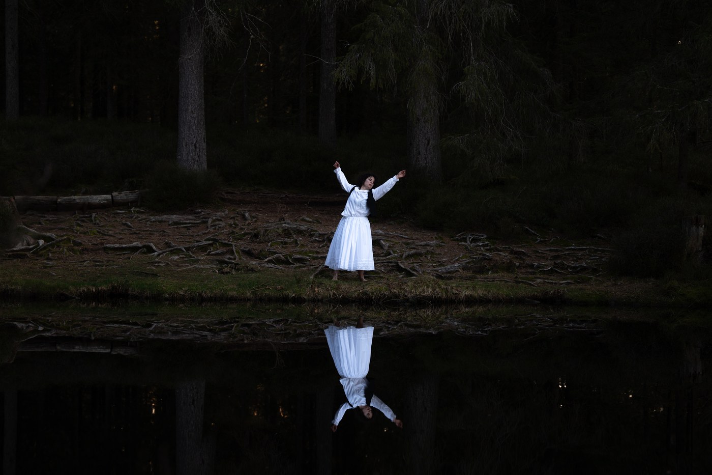 Une femme en robe blanche, bras levés, au bord d’un étang forestier sombre où elle se reflète